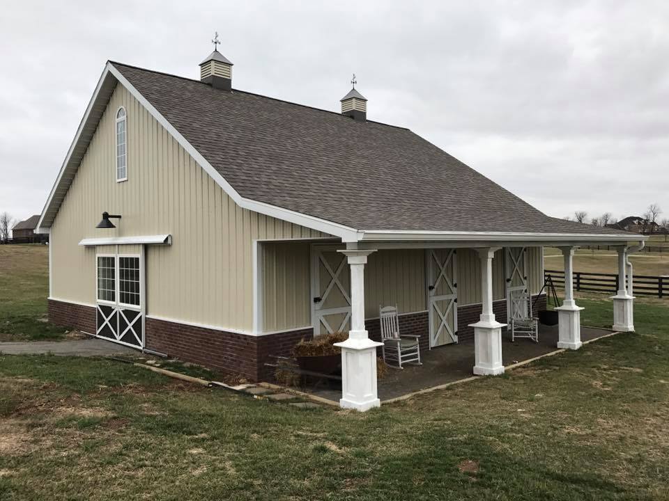 A tan horse stable with a porch and rocking chairs. It has a brown roof, white trim, and brick at the base.