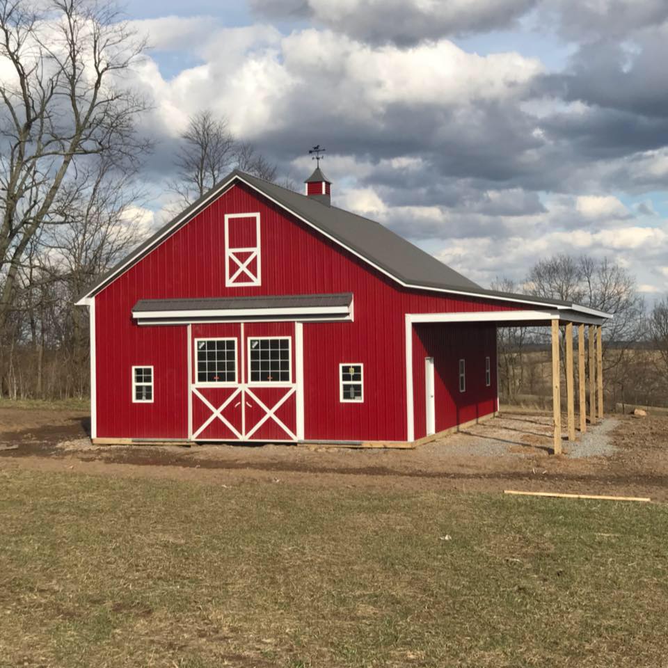 Red barn with white trim and a gray roof, under a cloudy sky. It has a small covered porch and a weathervane.