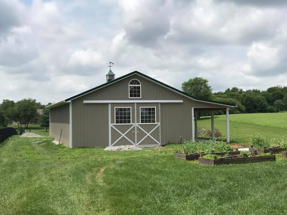 A gray barn with a green roof and white trim sits in a grassy field on a cloudy day. A small garden is in front.