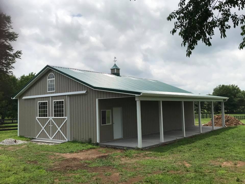 A gray barn with a green roof and an open-air porch.  Features a window, white trim, and a weathervane in a grassy field.