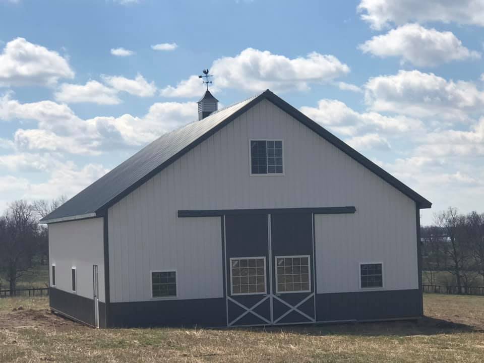 White and blue barn in a field under a partly cloudy sky. Barn has a black roof and accents, with a weathervane on top.