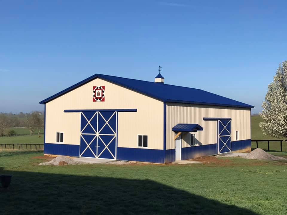 A tan and blue barn with a quilt block decoration on its front. It has large sliding doors and sits on a grassy field.