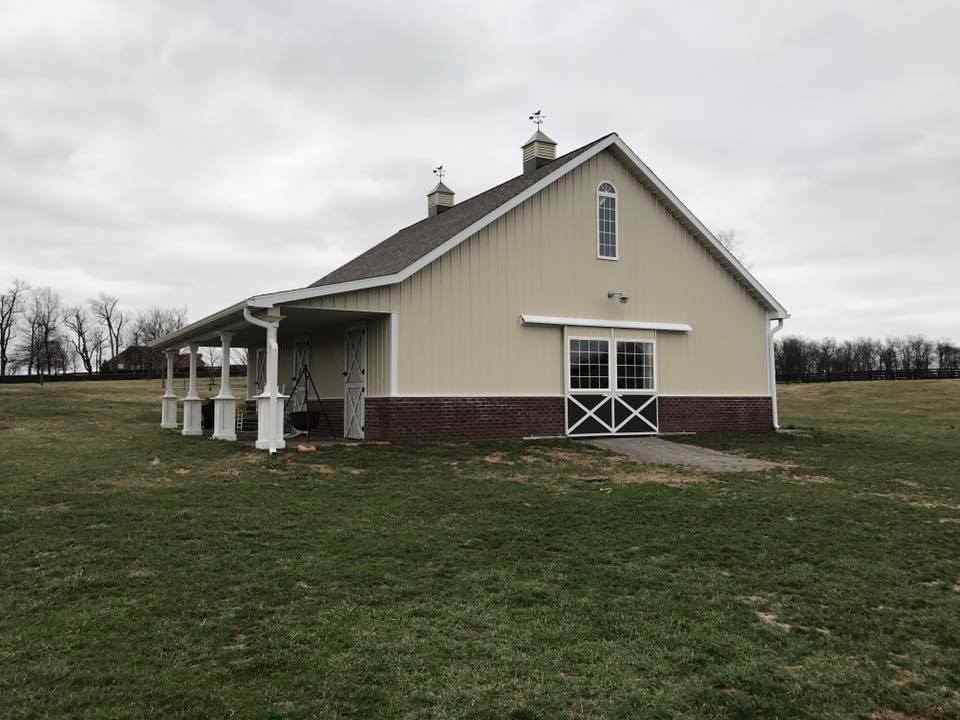 Tan barn with brick base, porch, and green grass field under an overcast sky.