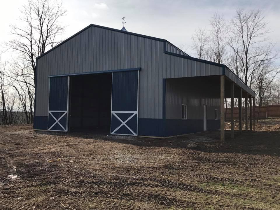 Blue and gray metal barn with large sliding doors and a covered side porch, set in a dirt lot with bare trees in the background.