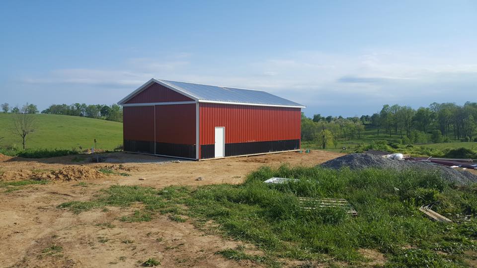 Red and black barn with a silver roof in a rural setting, with green grass and trees under a blue sky.