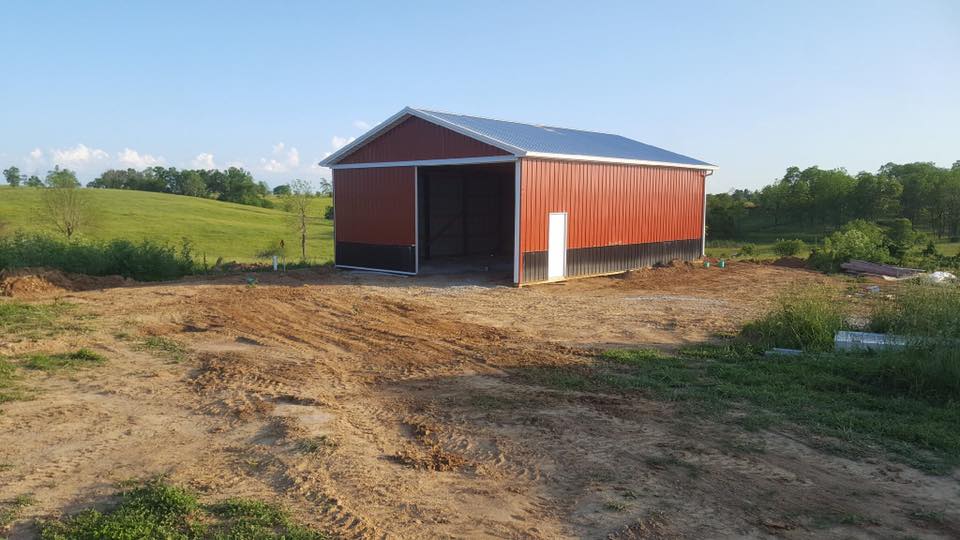 A red barn with a silver roof stands on a dirt patch, with a green field and trees in the background under a blue sky.