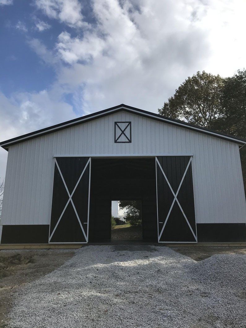 White and dark-trimmed barn with open doors, gravel path leading to a distant clearing, against a partly cloudy sky.
