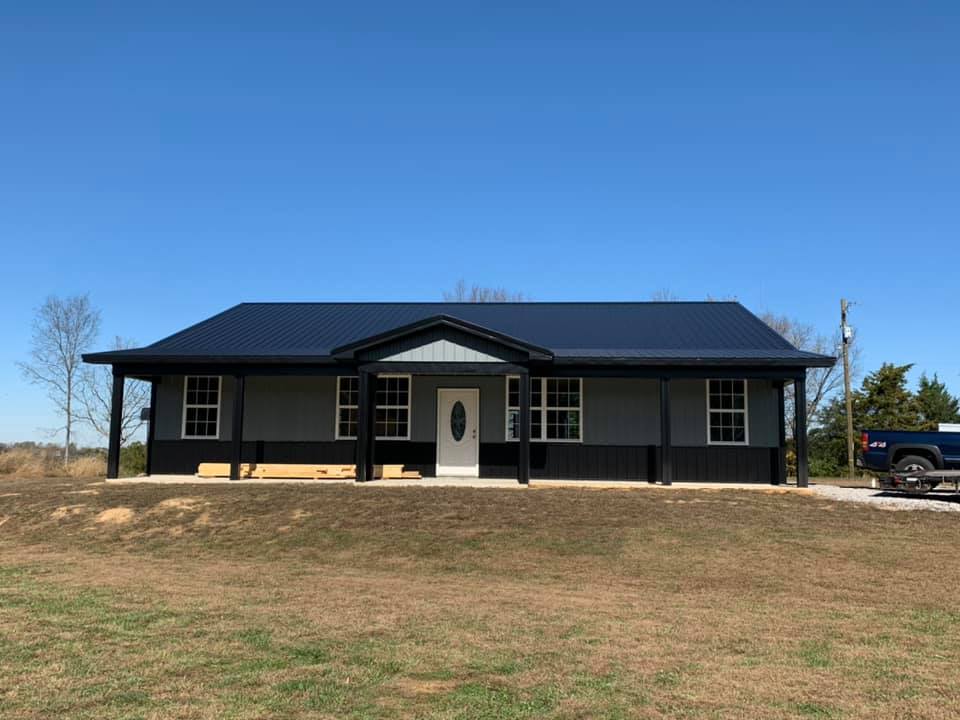 A gray and black ranch-style house with a blue roof and porch, set in a field under a clear blue sky.