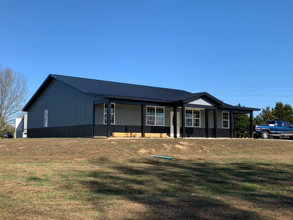 A blue and black metal building with a porch on a grassy hill. A blue truck is parked to the right.