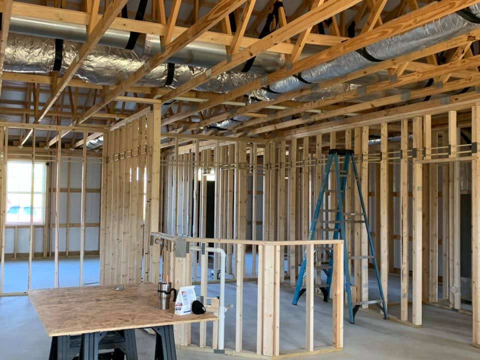 Interior framing of a house under construction; wood studs define walls and ceilings, with a ladder, ductwork, and tools visible.