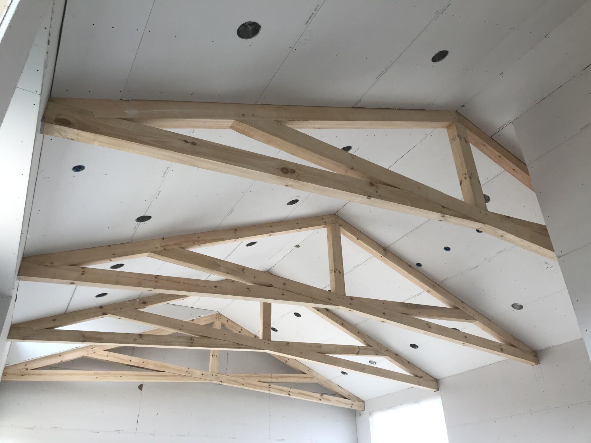 Wooden roof trusses are installed on a white ceiling, with recessed lighting. The room's walls are also white.