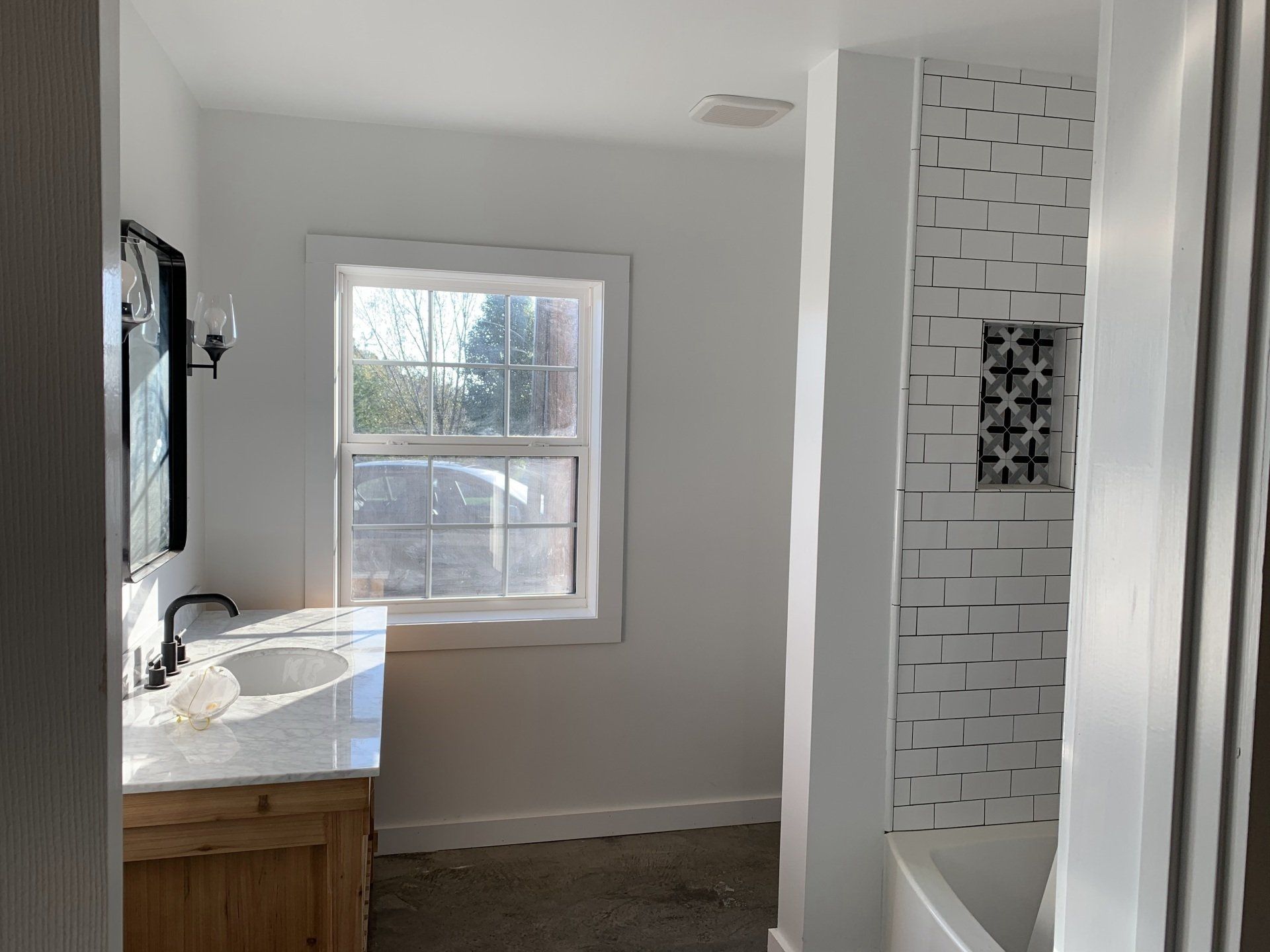 A white bathroom with a vanity, a window, and a tiled shower wall. The vanity has a wooden cabinet and a gray countertop.