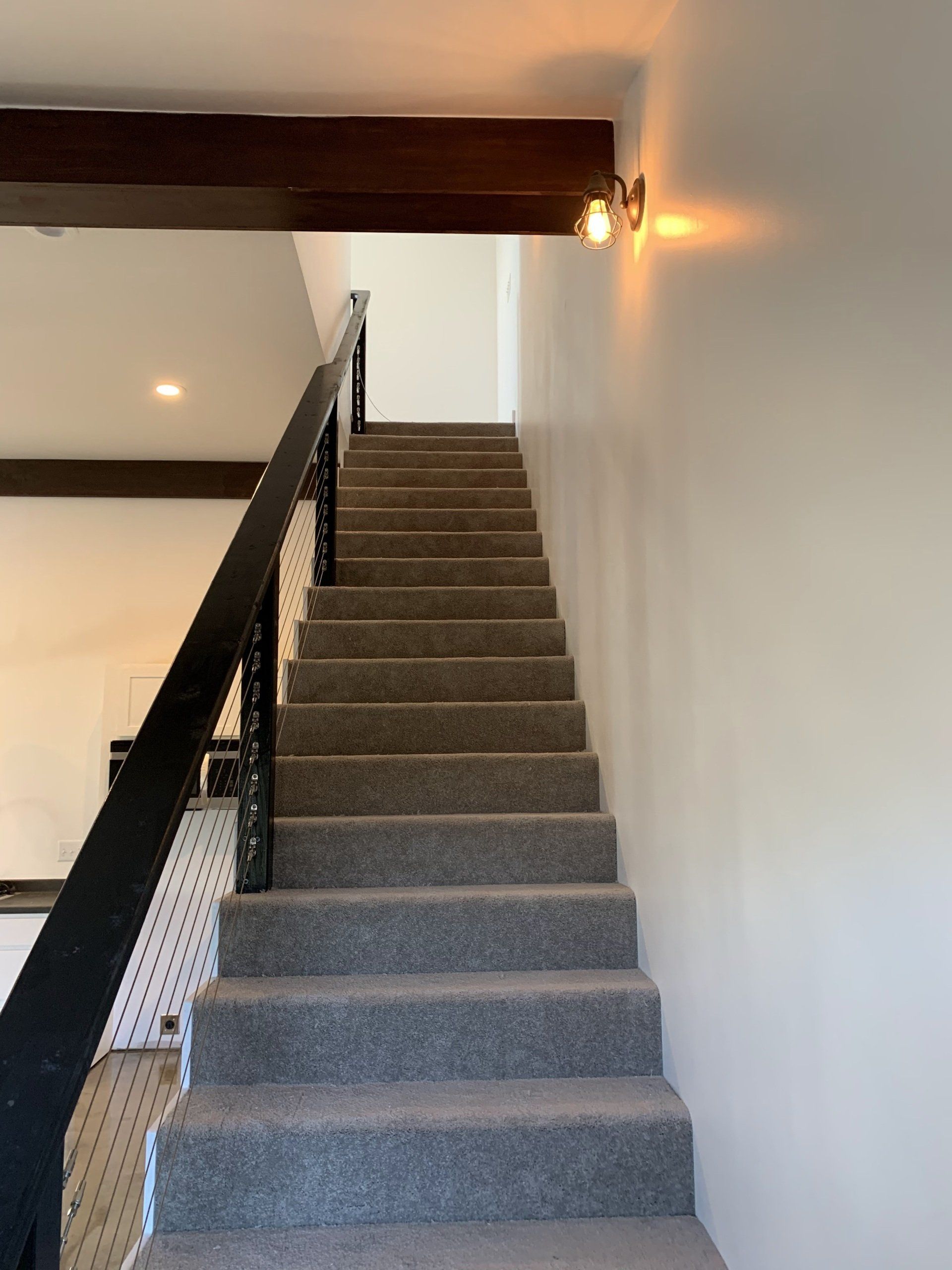 Staircase with gray carpet and a black railing ascending to a white wall, illuminated by a warm light fixture.