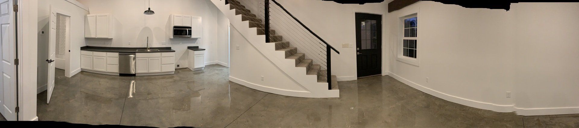 A modern interior with white walls, a kitchen with a black countertop, a staircase, and a dark door. The floor is concrete.