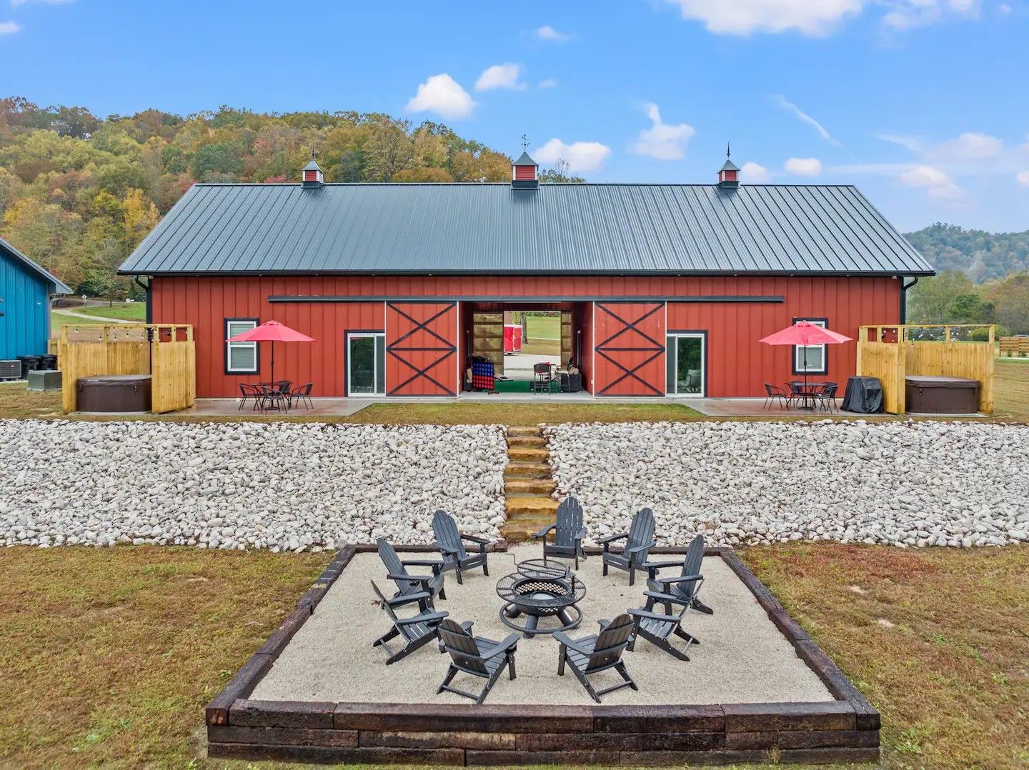 Red barn with open doors, flanked by patios and a fire pit seating area. Scenic outdoor setting.