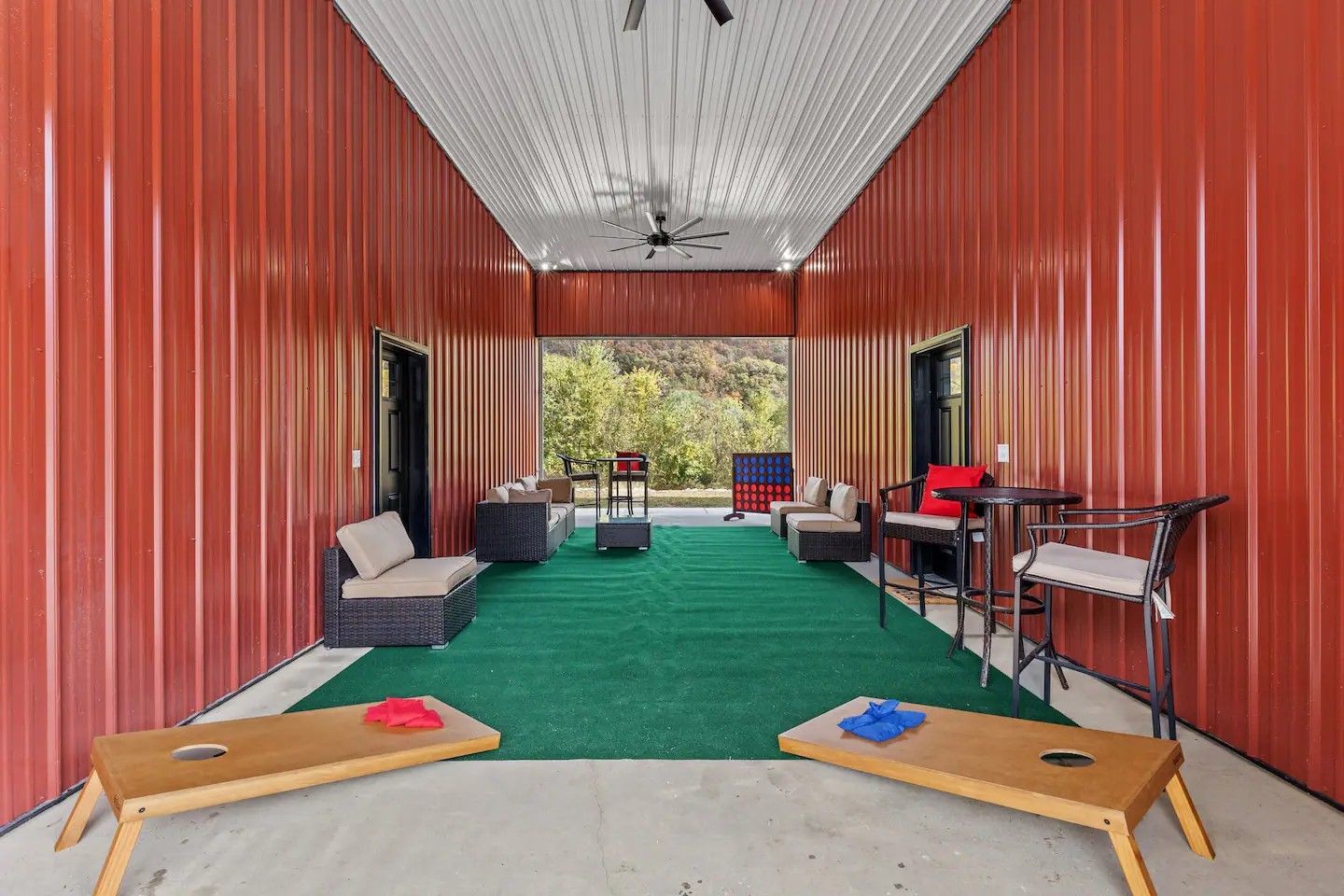 Open-air game room with red corrugated walls, green turf floor, and cornhole boards. Furnishings include seating and tables overlooking a wooded view.
