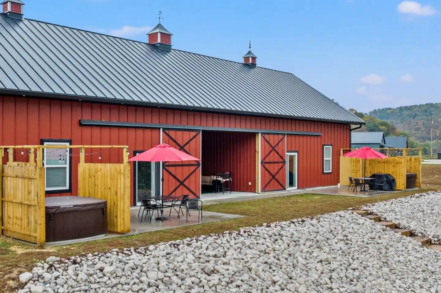 Red barn-style building with open sliding doors, outdoor seating with red umbrellas, and a hot tub.