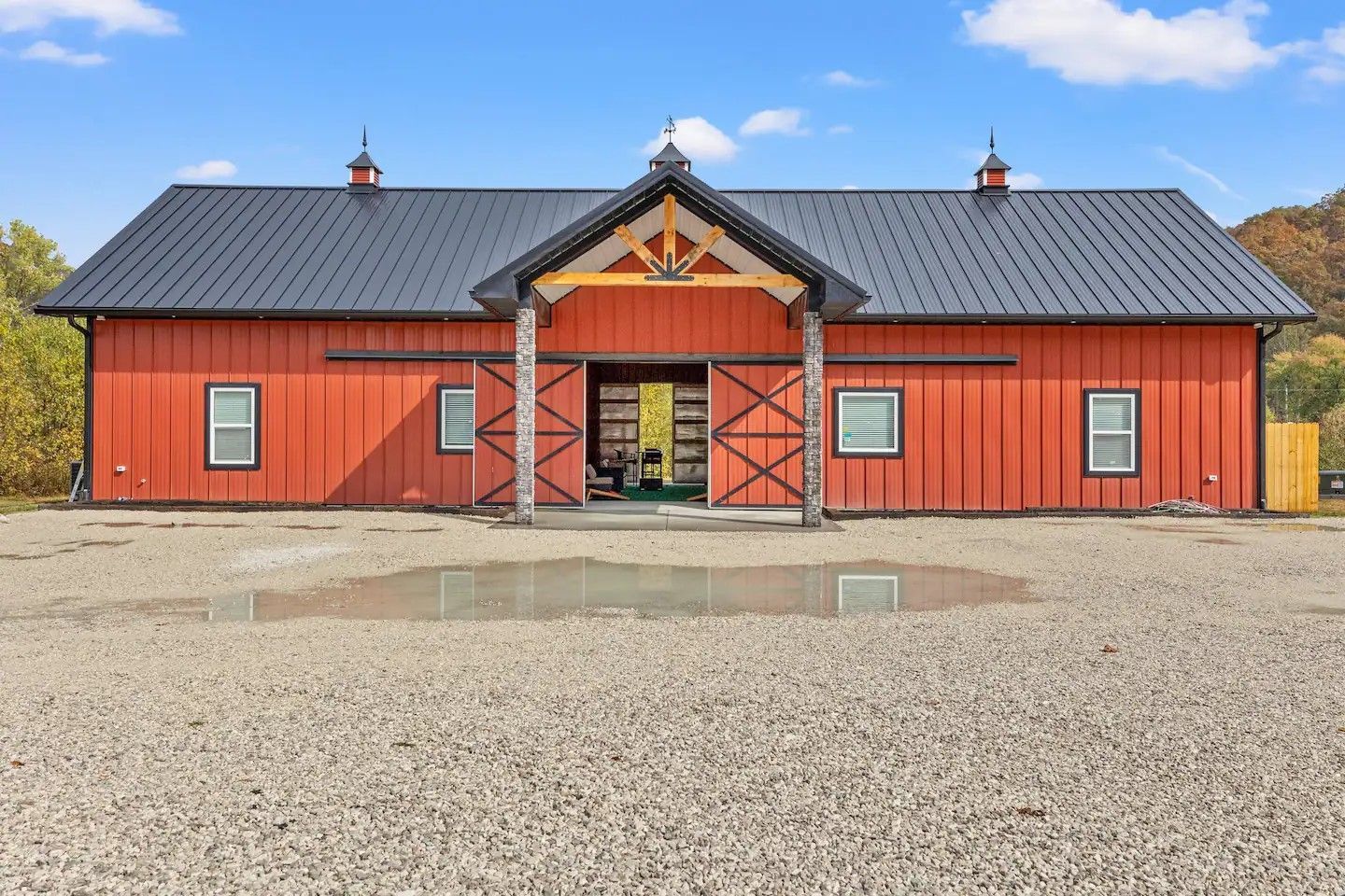Red barn-style building with black roof, white windows, and an open doorway. The setting is a gravel yard with a small puddle.