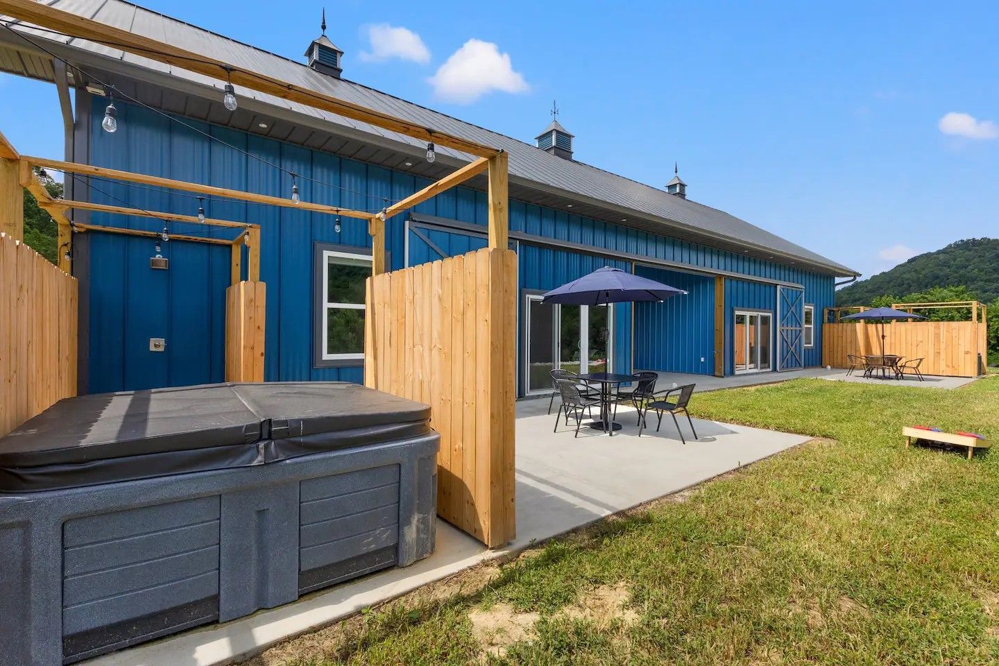 A blue barn with a hot tub, patio furniture, and yard on a sunny day. Wooden privacy screens are beside the hot tub.