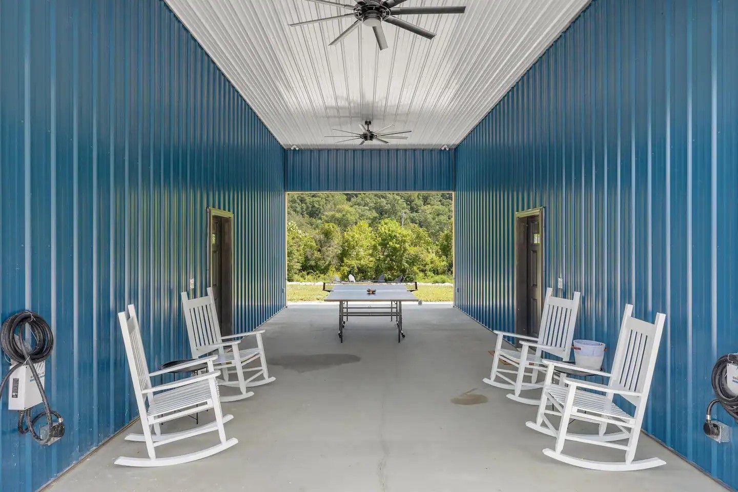 Blue corrugated metal porch with white rocking chairs, a foosball table, and an open view of trees.