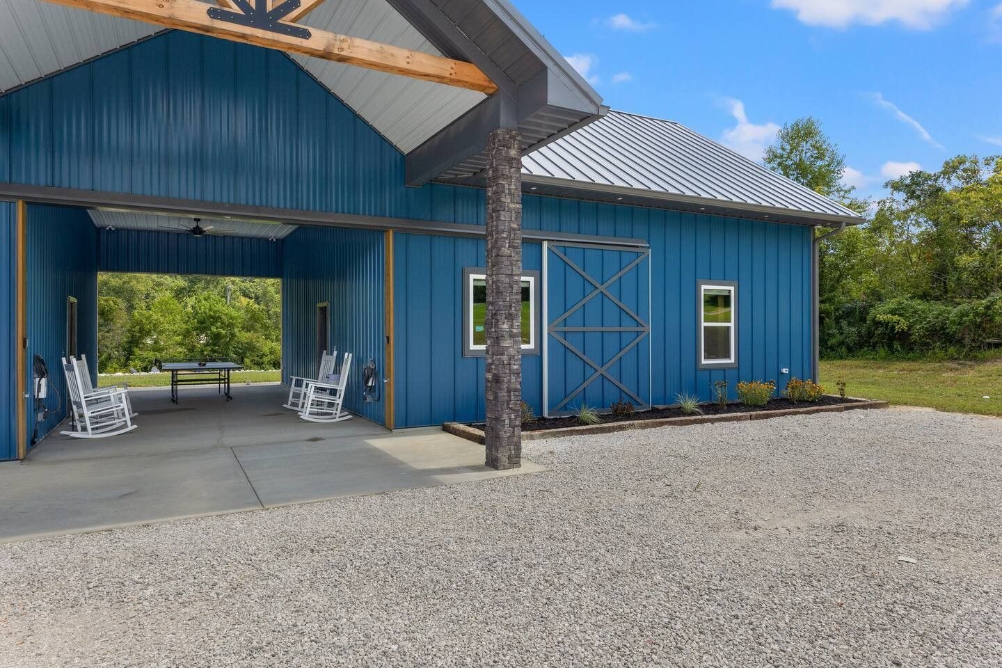 Blue barn with a covered patio featuring rocking chairs, with gravel driveway and a grassy field backdrop.