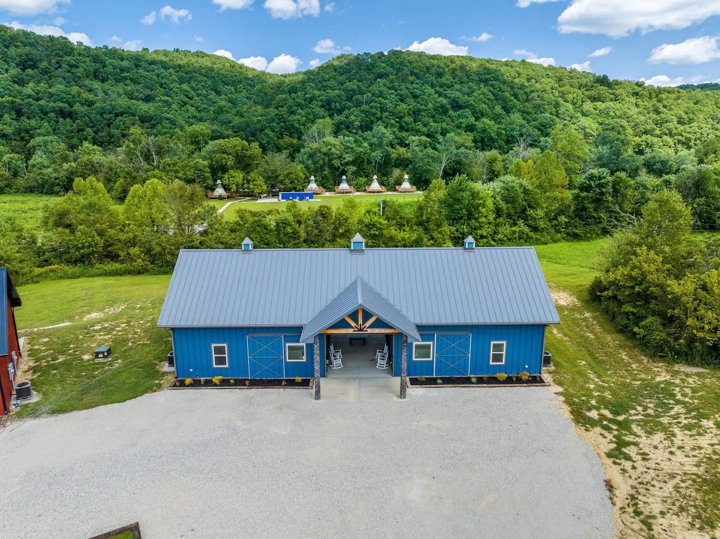 Blue barn-style building with a gravel driveway, surrounded by trees and a hilly landscape under a blue sky.