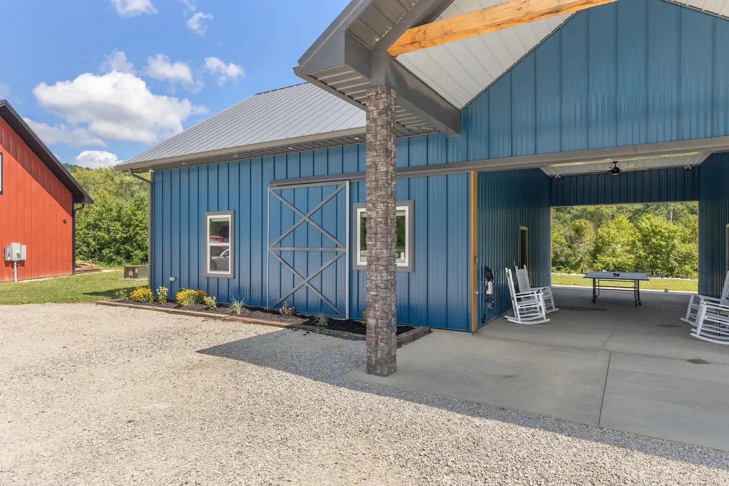 Blue barn-style building with a covered porch. A red building is visible to the left. The ground is covered in gravel.