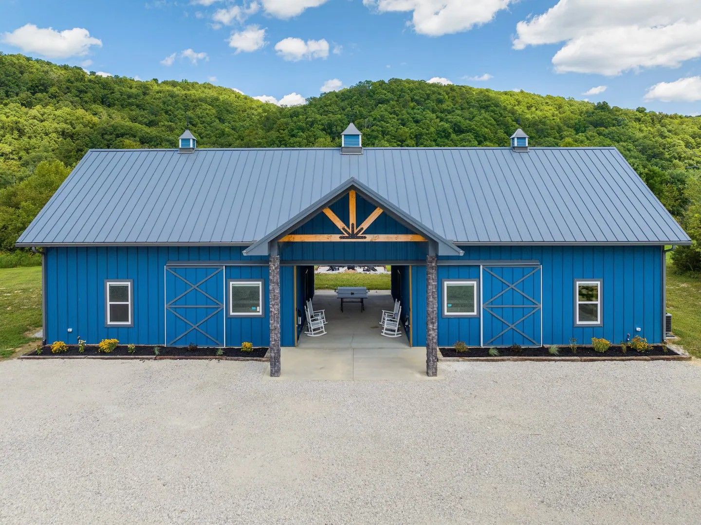 Blue barn-style building with a gray metal roof, set against a backdrop of green hills and a bright blue sky.