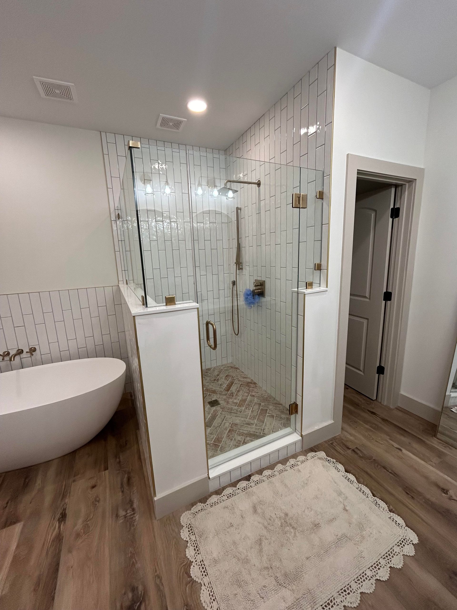 Modern bathroom with a glass shower, white tub, and wood-look flooring. The shower has pebble flooring.