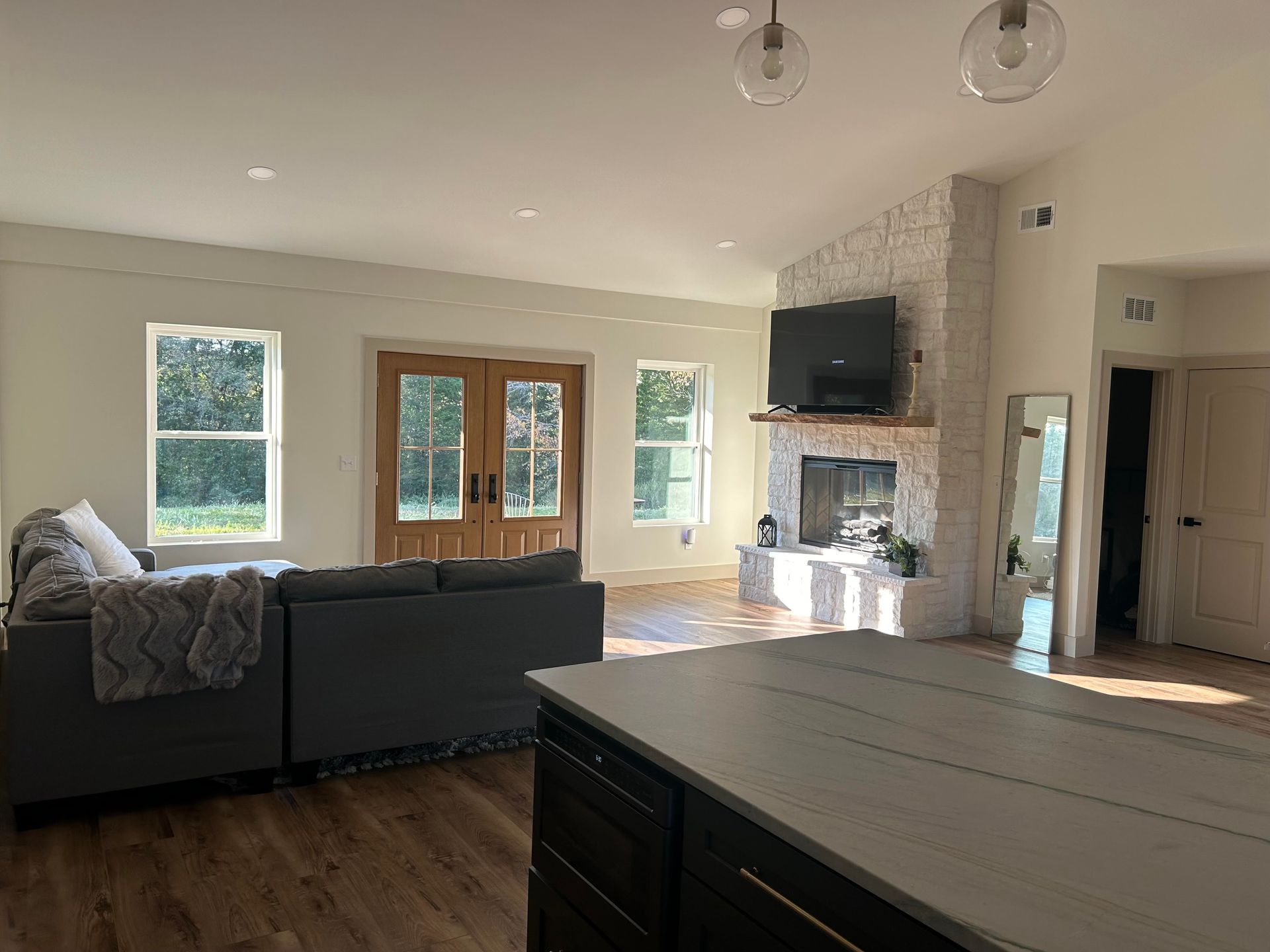 Spacious living room with a fireplace, large windows, and a kitchen island in the foreground. A gray sectional sofa sits near the windows.