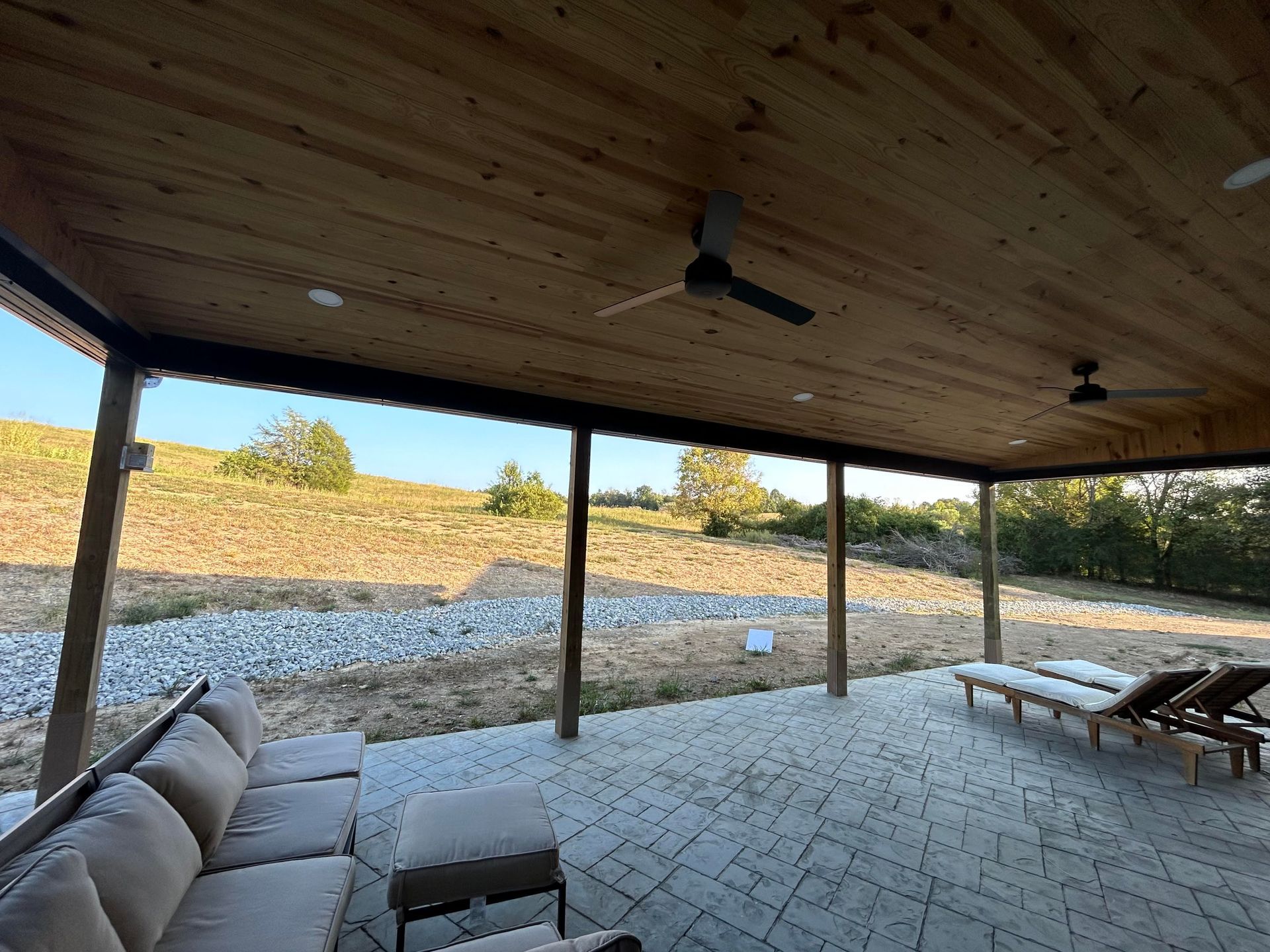 Covered patio overlooking a dry field. Beige couch and lounge chairs sit on a brick patio under a wooden ceiling with fans.