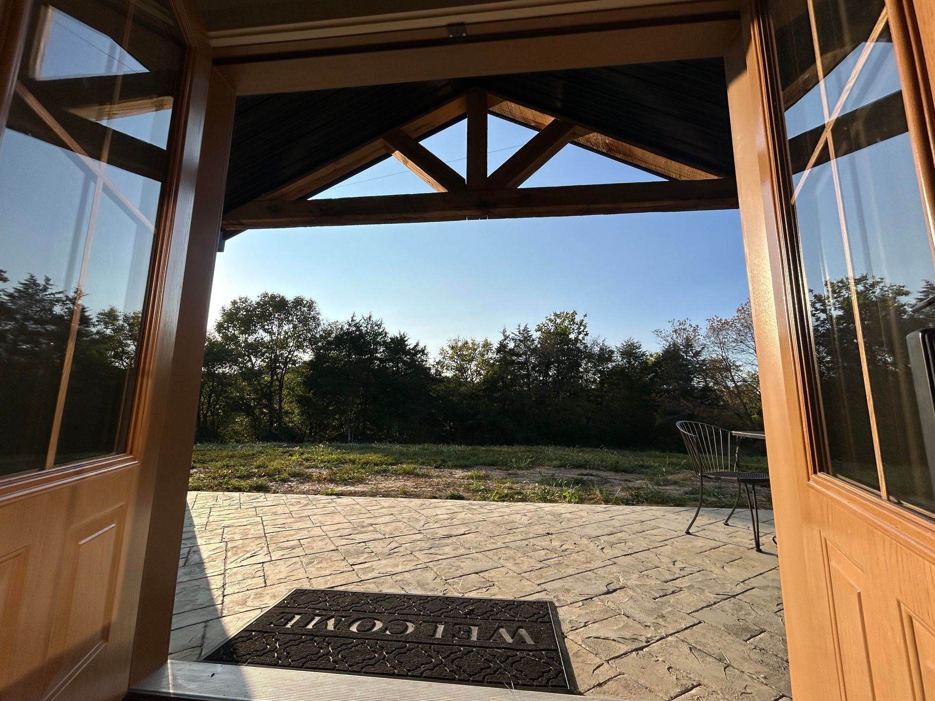 Open wooden doors lead to a patio with a welcome mat, a trellised roof, and a view of a grassy area and trees under a clear blue sky.