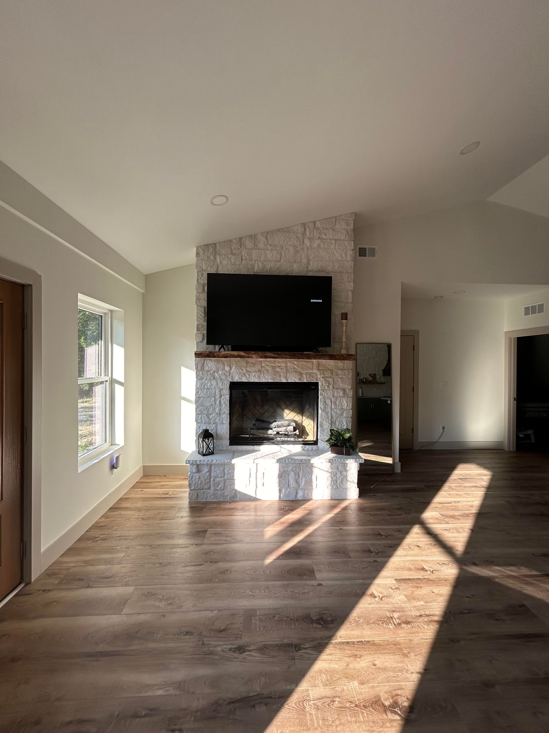 Spacious living room with hardwood floors and a stone fireplace, sunlight streaming through the window.