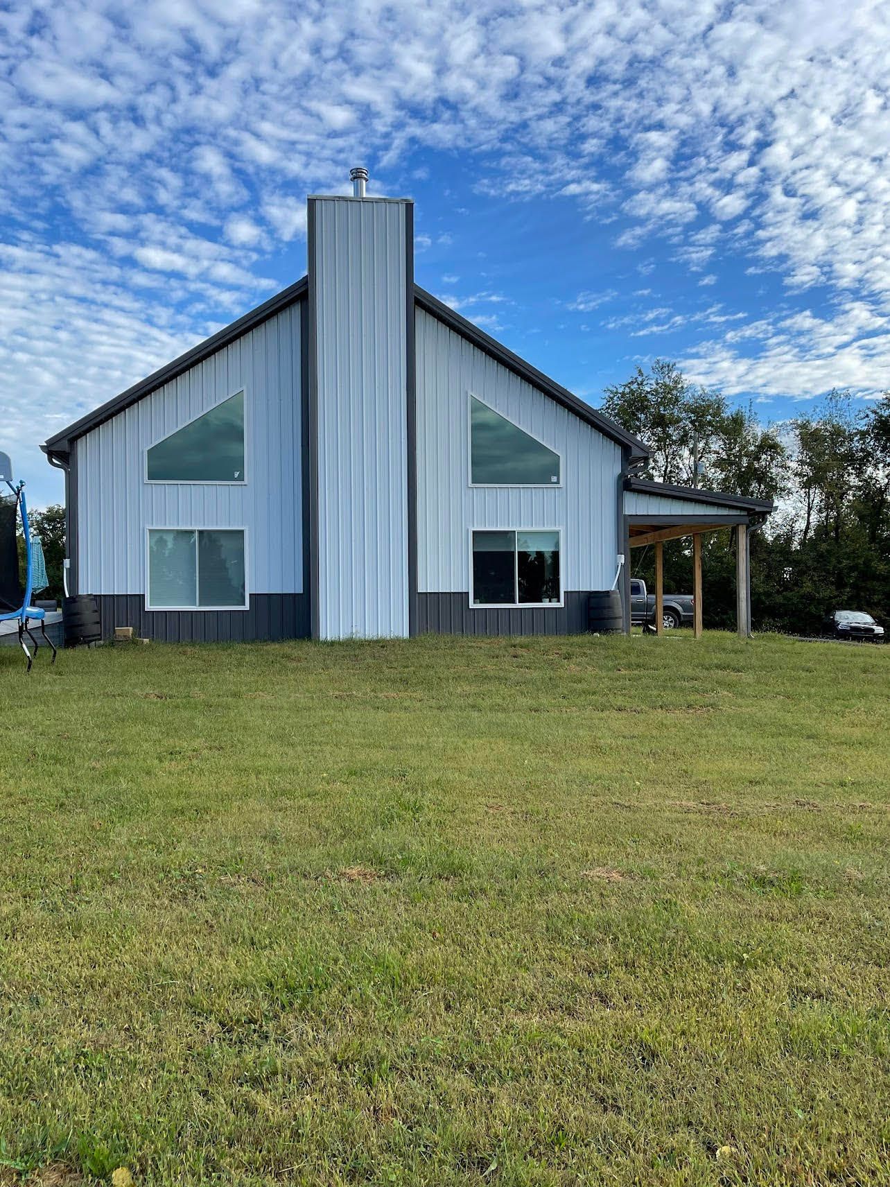 A light blue metal building with a tall chimney and large windows sits on a grassy hill under a cloudy blue sky.