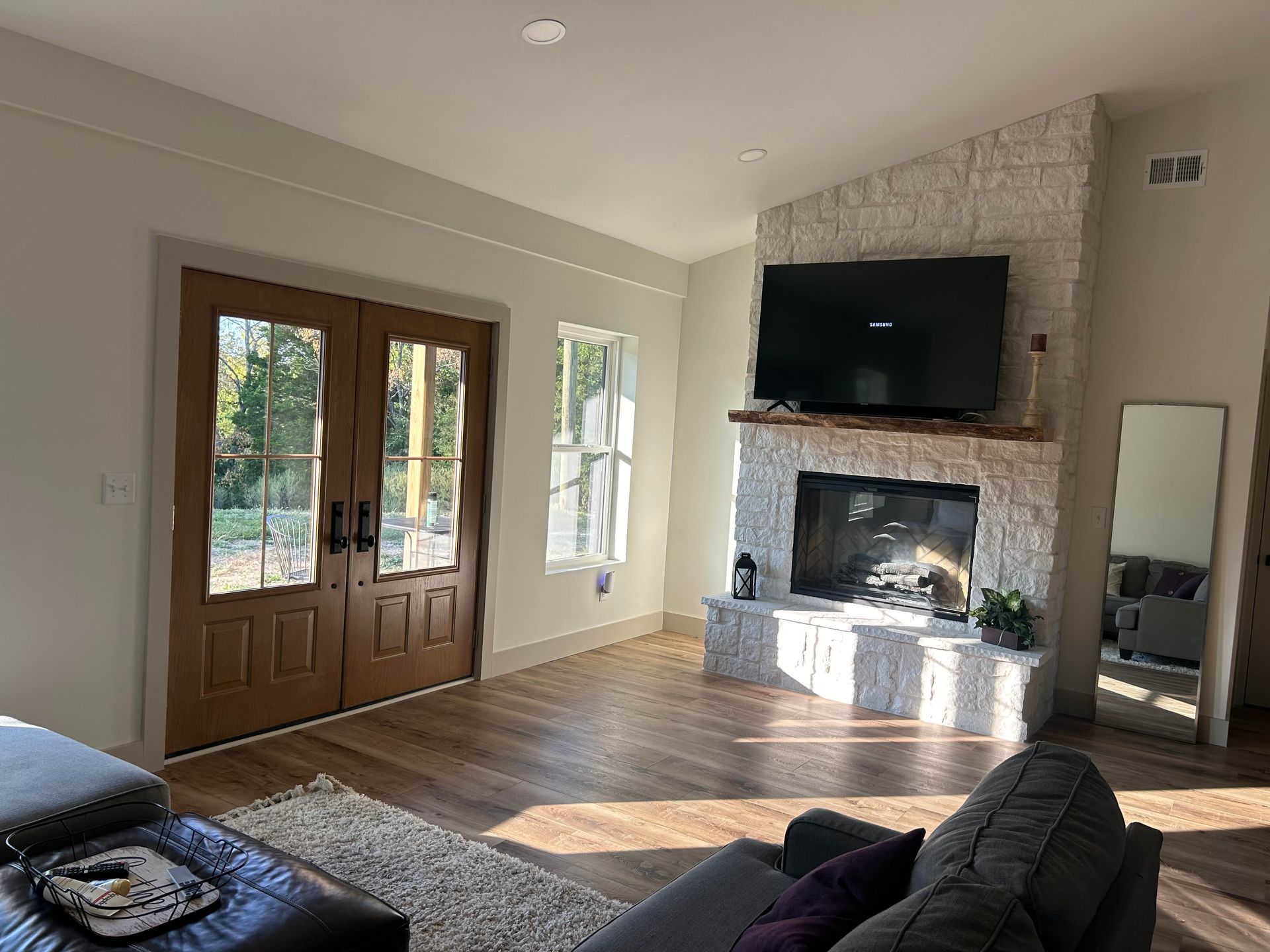 Living room with a fireplace, double doors, and a large TV. Sunlight streams in, illuminating the wood floors and stone accents.