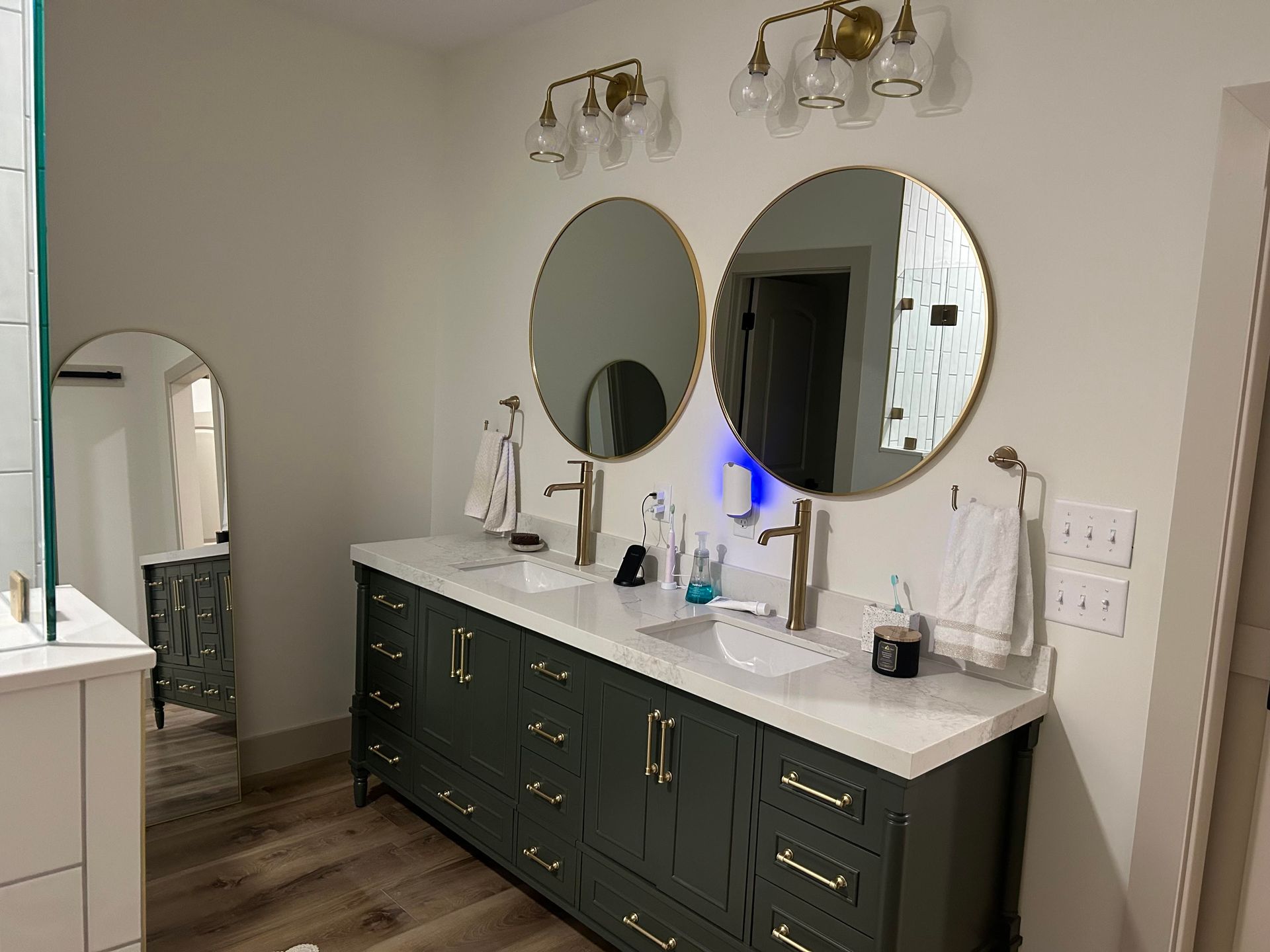A modern bathroom with a green vanity, two round mirrors, and gold fixtures.  There's a full-length mirror on the left side.