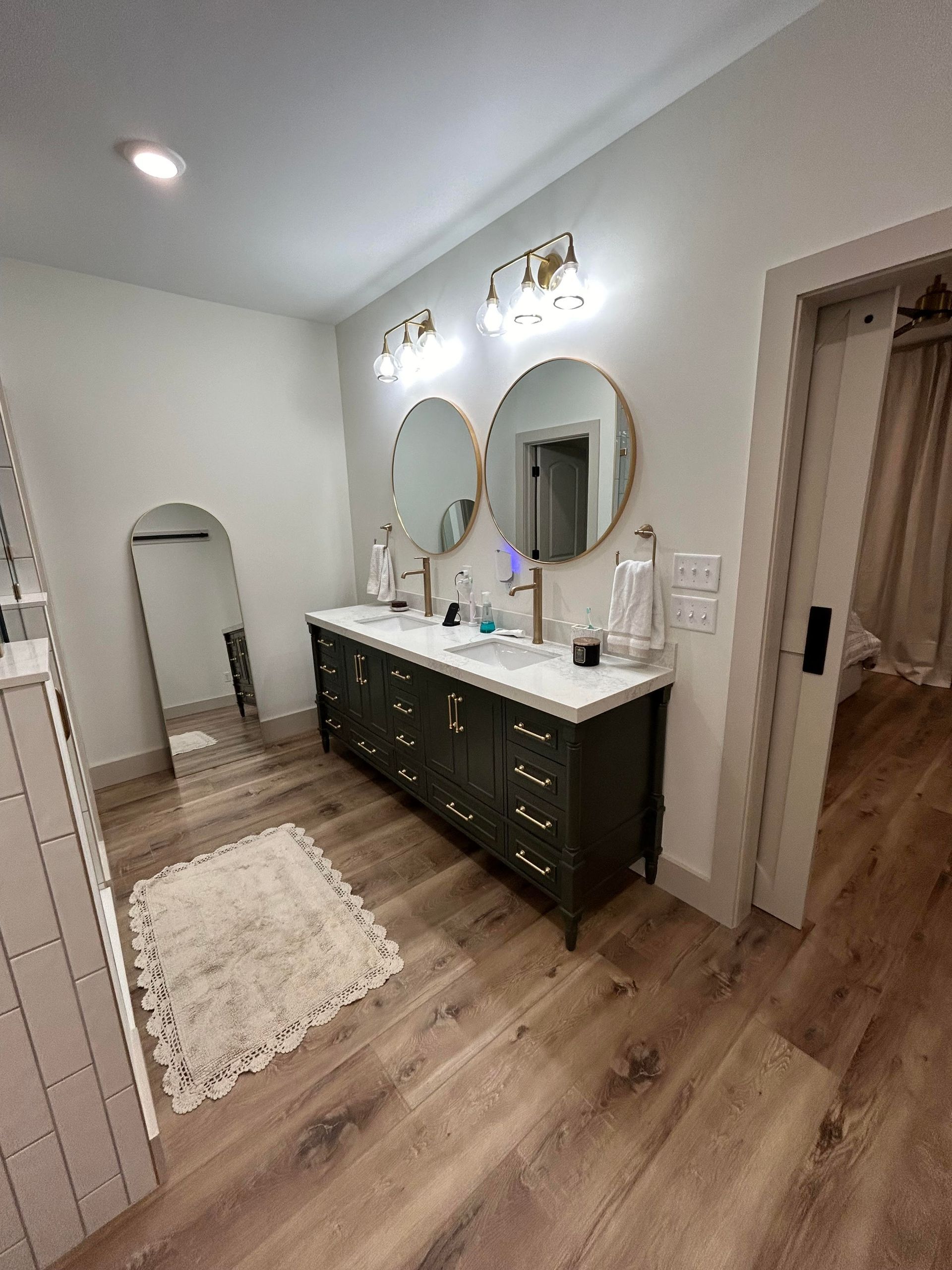 A modern bathroom with a dark green double vanity, two round mirrors, and light wooden floors. A doorway is on the right.