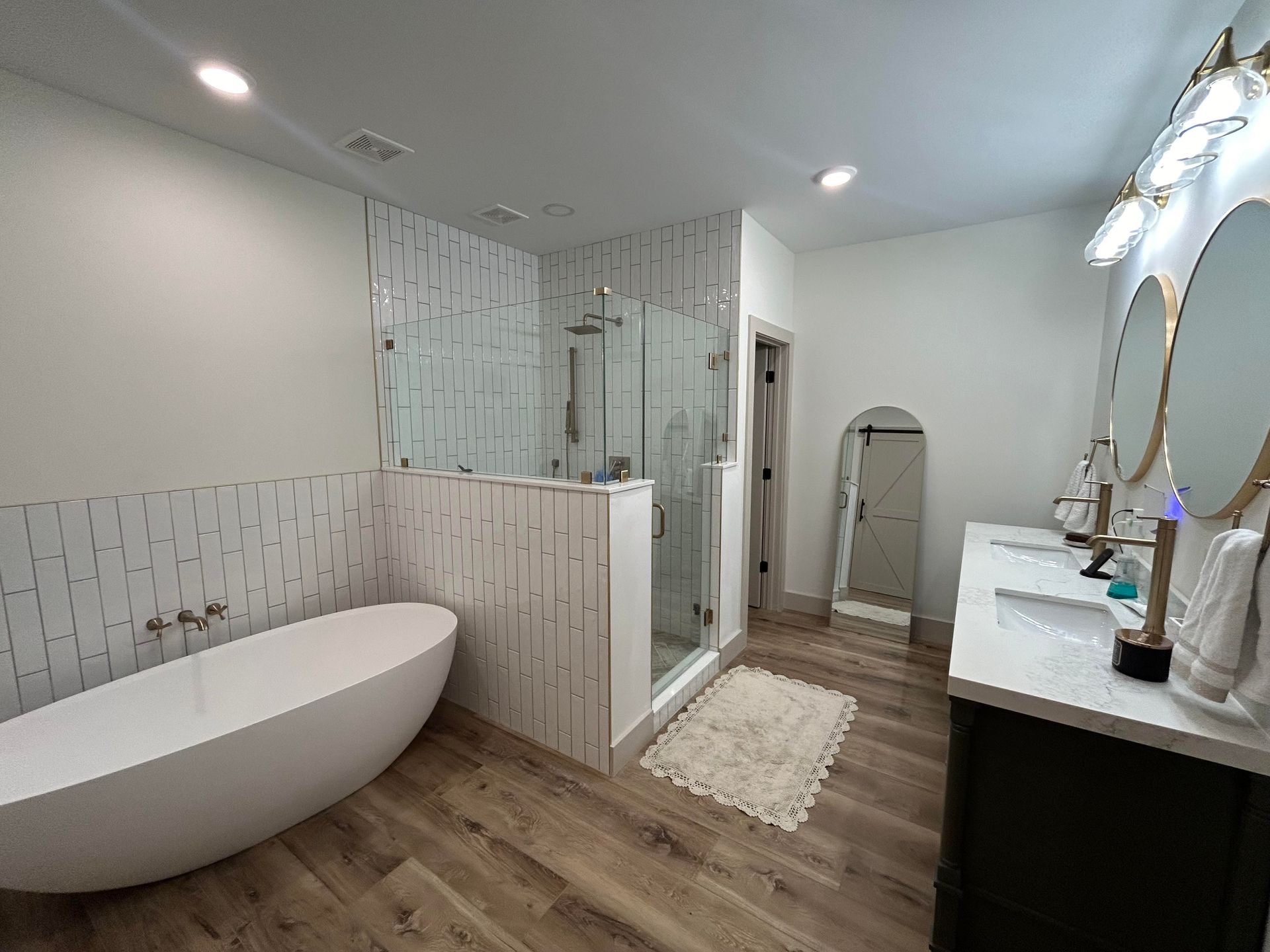 Modern bathroom with a white tub, glass shower, and double vanity with gold mirrors. The floor is wood-look tile.