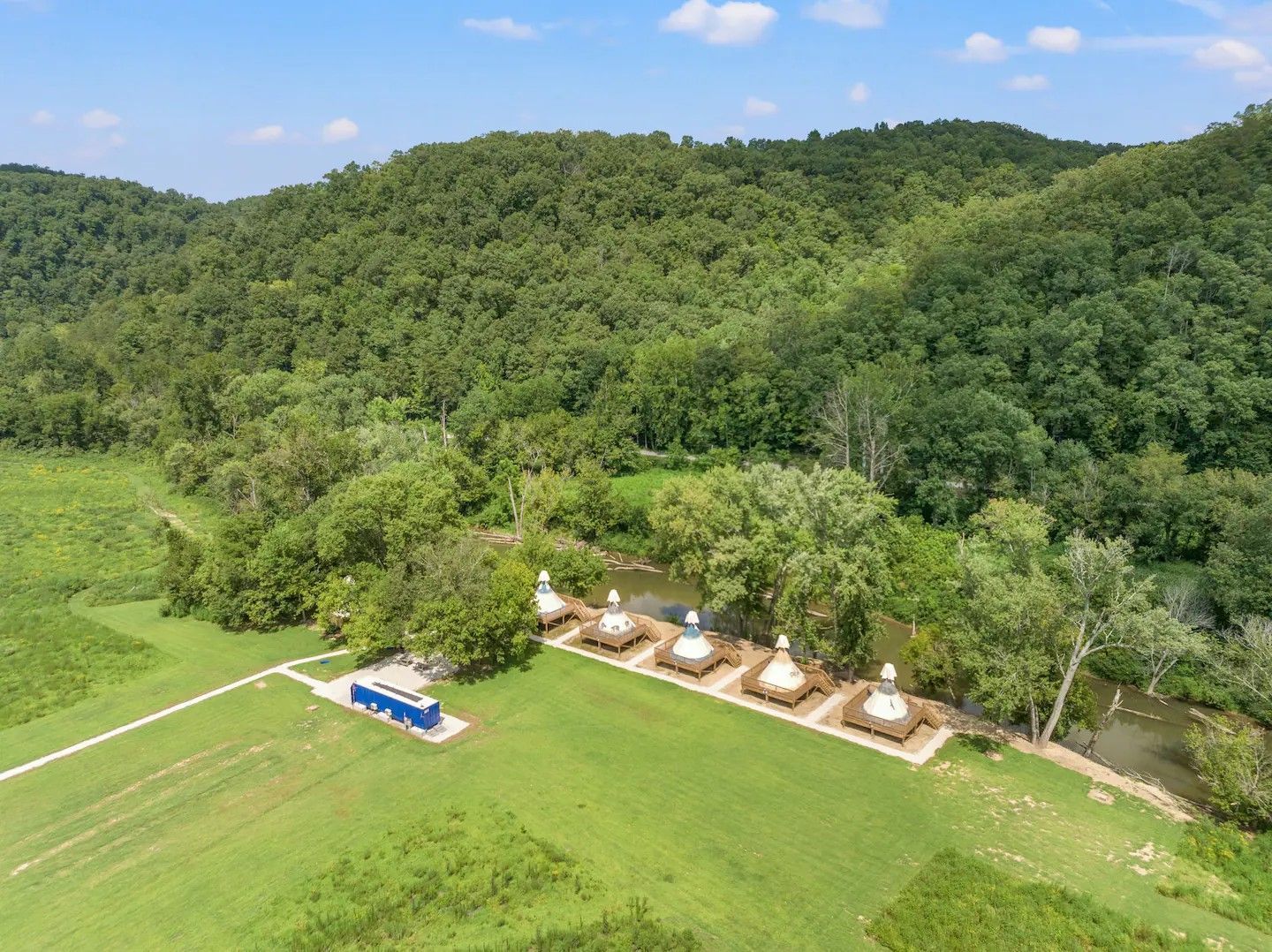 Aerial view of white structures with conical roofs along a river, surrounded by green fields and a forested hillside.