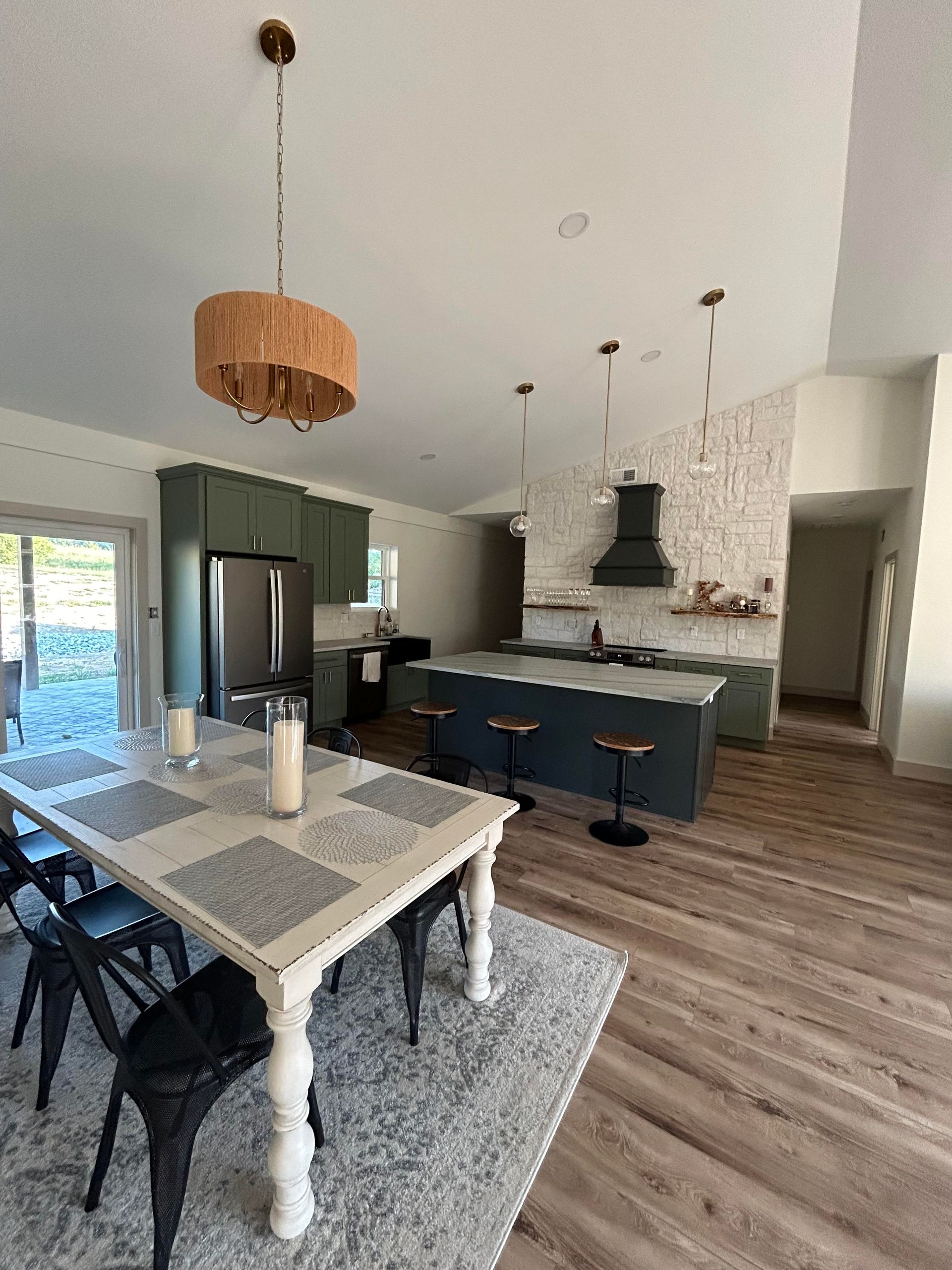 Open-concept kitchen and dining area. Features green cabinetry, a blue island, white brick accent wall, and a farmhouse-style dining table with black chairs.