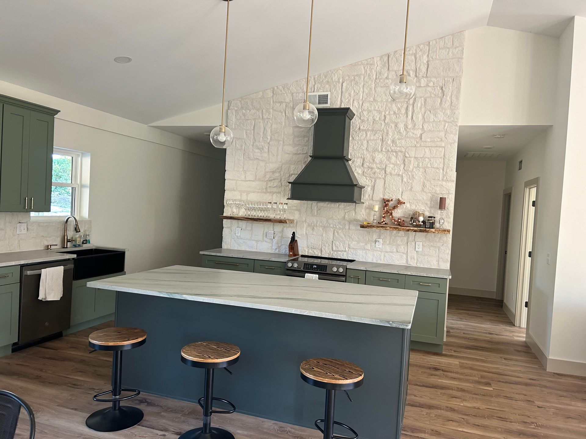 Modern kitchen with sage green cabinets, dark gray island, and brick backsplash. Three pendant lights hang over the island with stools.