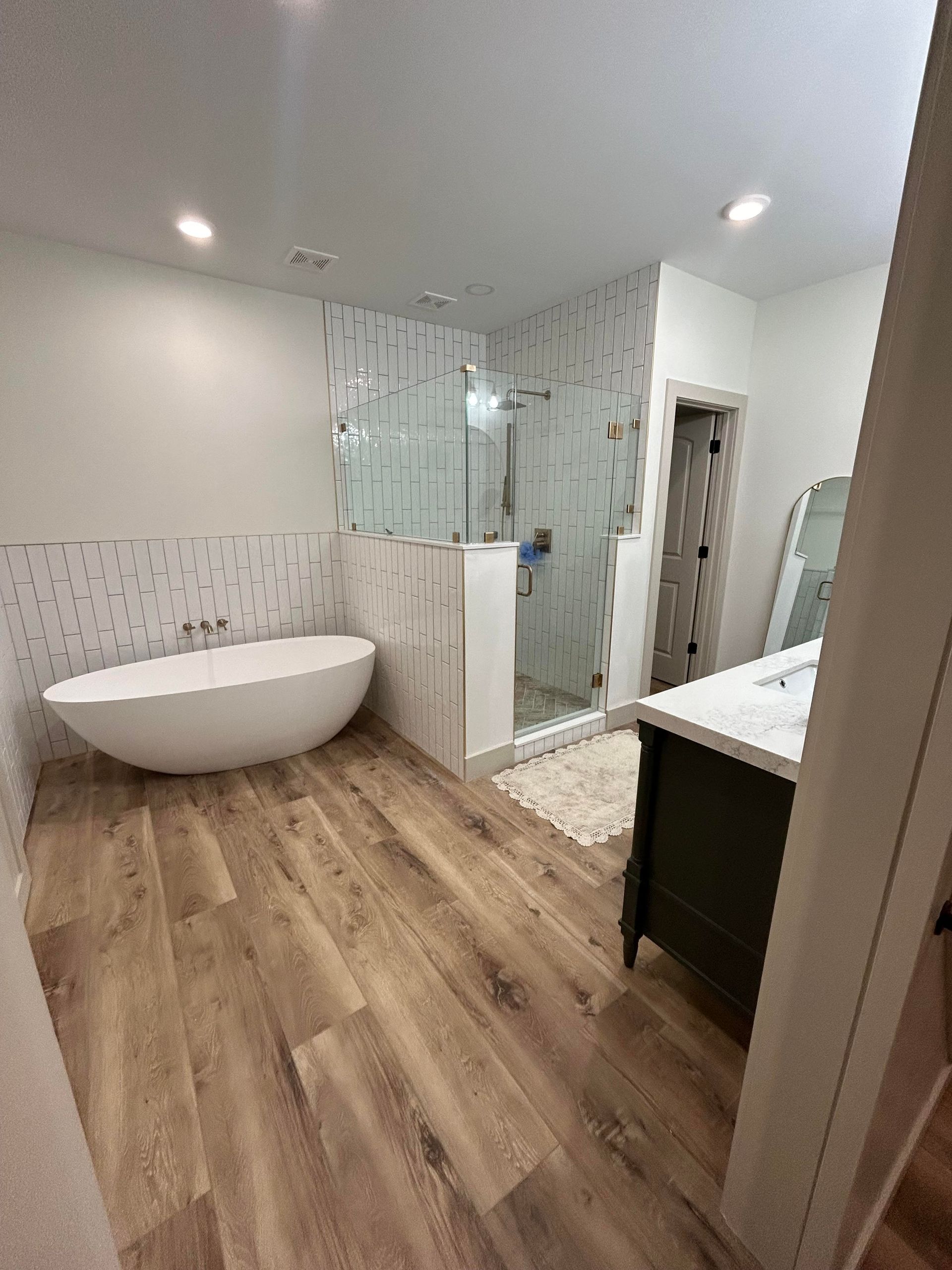 A modern bathroom with a freestanding tub, glass shower, and dark wood vanity with a stone countertop. Wooden floors and white walls.