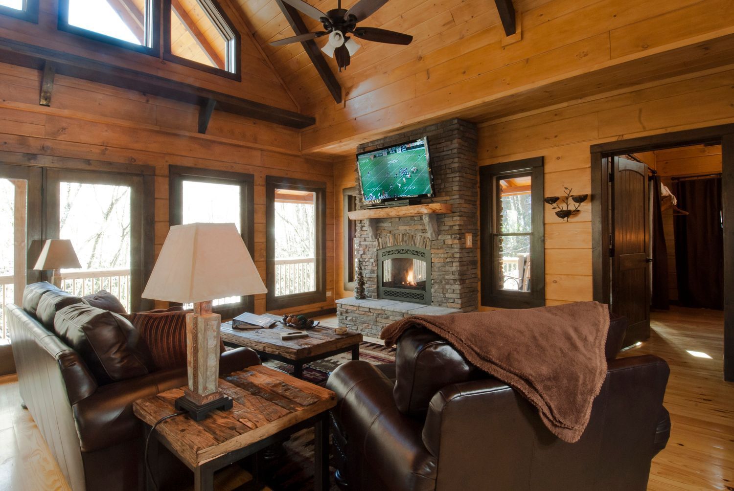 Cozy cabin living room with leather furniture, stone fireplace, and large windows.