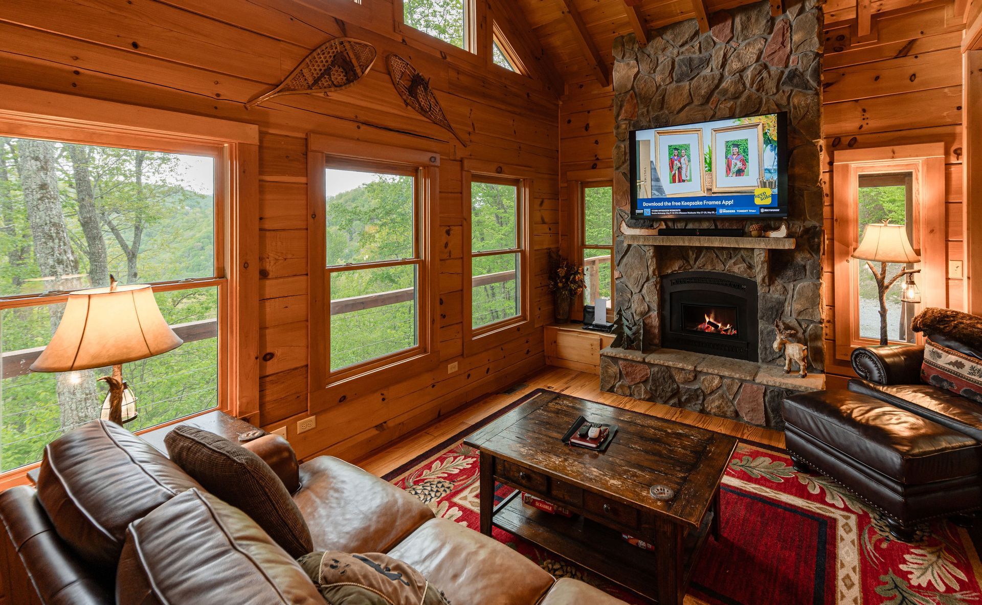 Cozy cabin living room with stone fireplace, TV, leather sofa, windows, and mountain views.