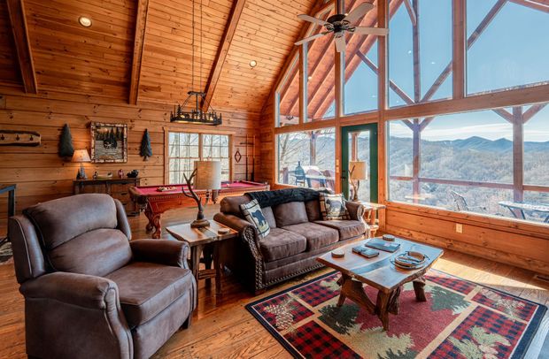 Cozy cabin interior with wood walls, large windows showcasing mountain views, and comfortable furniture.