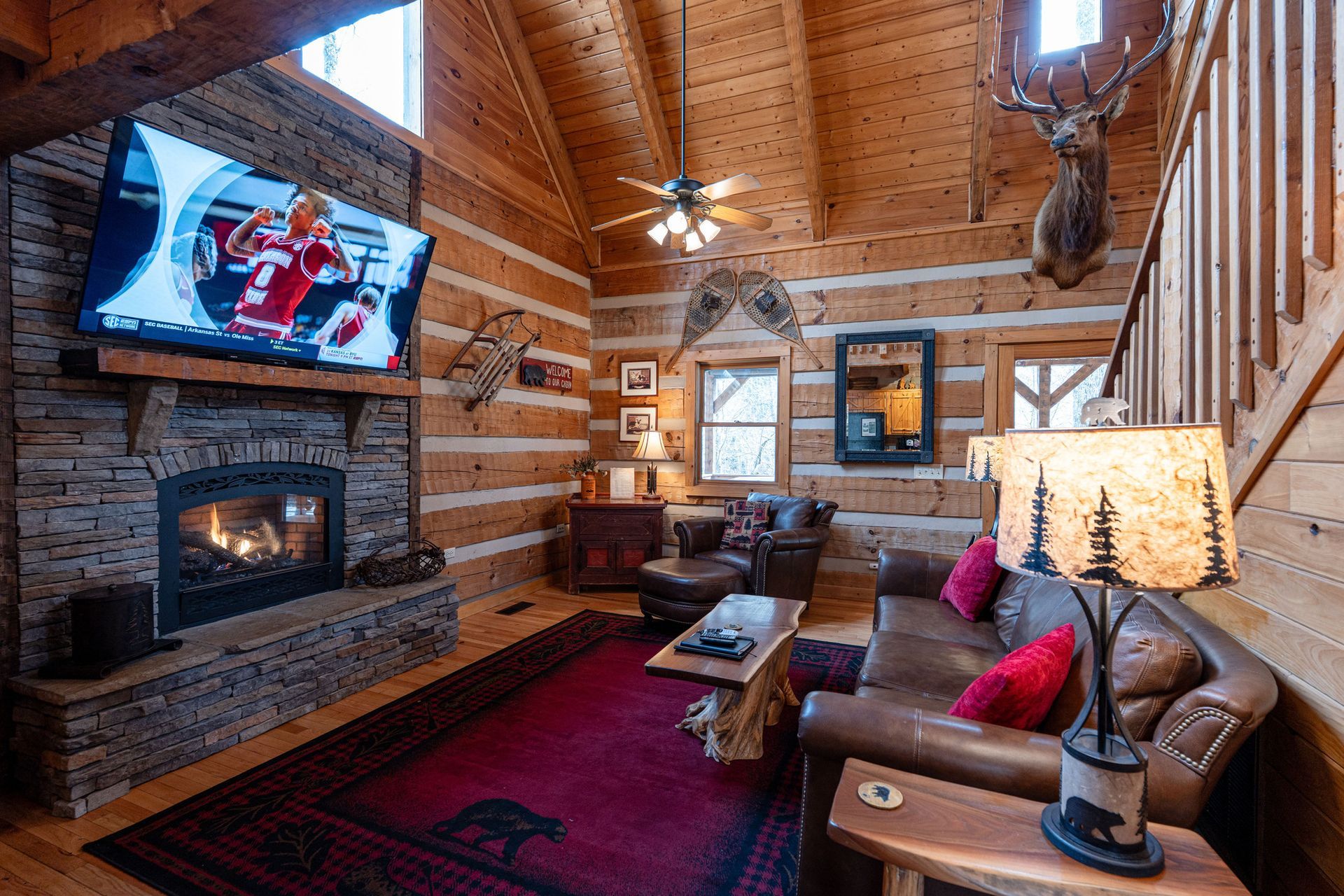 Cozy cabin living room: stone fireplace with TV, brown leather furniture, red rug, wooden walls, antler decor.