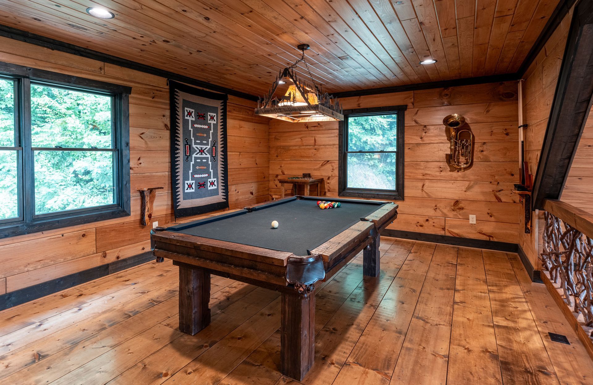 Pool table in a rustic wooden room with windows, art, and a decorative light fixture.