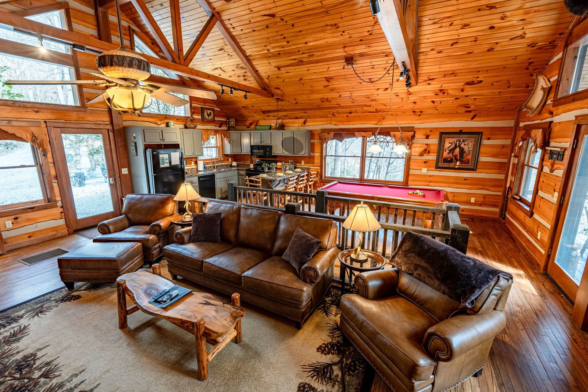 Cozy cabin interior with a living area, leather furniture, pool table, and kitchen. Wooden walls and ceilings.