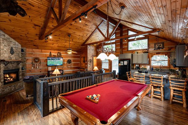 Rustic cabin interior with pool table, fireplace, and kitchen. Wooden beams and paneling; red pool table.