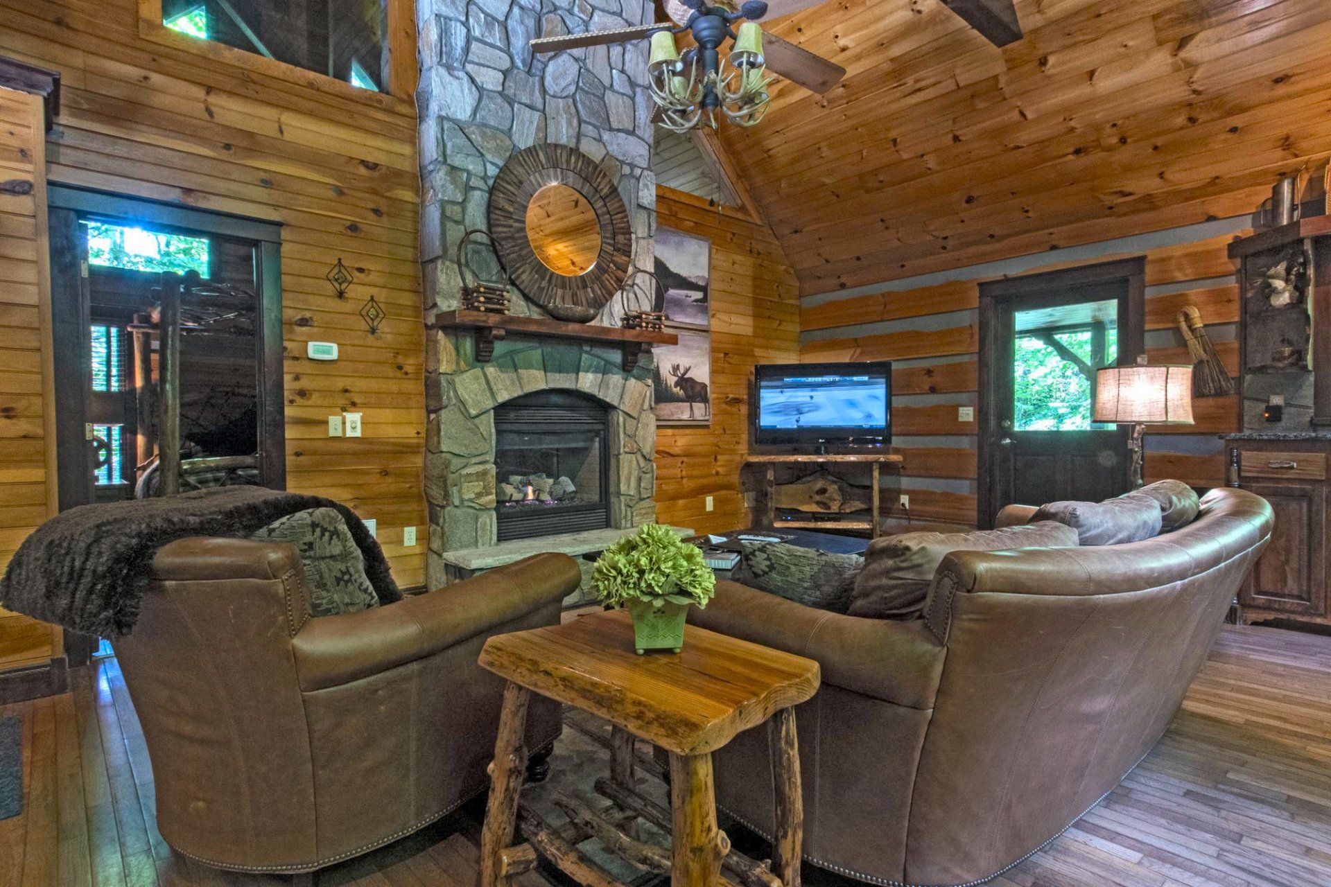 Cozy cabin living room with stone fireplace, leather furniture, TV, and wooden walls.